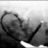 Black-and-white pixelated image of a person with glasses and dark hair, wearing a dark jacket over a light shirt, holding an object near their face.