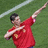 A person in a red sports jersey with yellow accents performs a celebratory gesture on a green field with white lines.