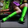 A static image shows a person in a bent-over pose wearing bright green thigh-high boots and a purple accessory, with green foliage in the background.