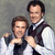 Two men in white shirts and dark vests pose together against a muted gray background, with one man's arm around the other's shoulder.