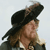 Pixelated image of a person with long dark hair wearing a black tricorn hat, light-colored shirt, and dark jacket against a light background.