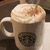 A white ceramic mug with a dark circular logo on its side, filled with light brown liquid topped by frothy foam and sprinkled with cinnamon or cocoa powder.