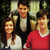 Three people stand together in a pixelated image; two wear white shirts, one wears red with white trim. Background is dark and indistinct.