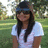 A static pixelated image of a person with long dark hair wearing a light blue shirt and dark cap, standing outdoors with greenery in the background.