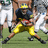Pixelated image of a football player in yellow pants and dark jersey holding a ball, with other players visible on a green field background.