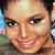 Pixelated close-up of a smiling person with dark hair, light skin, and visible teeth, set against a blurred background with blue and green tones.