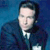 Pixelated image of a person in a dark suit with a white shirt and dark tie against a blue background.