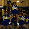 Cheerleaders in blue and yellow uniforms pose with arms raised, one standing on others' shoulders in a gymnasium setting.