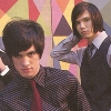 Two individuals in formal attire pose against a colorful geometric background with triangular shapes in red, yellow, and purple.