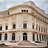 Static image of a classical building facade with columns, multiple windows, and a curved roofline under a light sky.