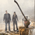Static 50x50 pixel icon showing two figures in dark clothing standing on a beach with a large fishing rod and tackle box, rendered in low-resolution grayscale tones with brown/orange accents.