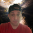 A person wearing a dark cap and red shirt appears in a pixelated static image with a blurred background containing light and dark areas.