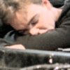 A person with light brown hair lies down with eyes closed, wearing a dark long-sleeve shirt, resting on a dark textured surface.