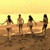 Four figures stand on a sandy beach at sunset, facing away from the viewer toward water under a yellow-orange sky.