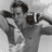 A black-and-white image of a shirtless man with dark hair holding two microphones near his face, standing against a light background.