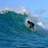 A surfer rides a large blue-green ocean wave under a light blue sky with scattered white clouds.