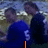 Two people in blue and dark shirts with white numbers on their backs, standing outdoors with text changing below them.