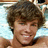 Pixelated close-up of a person with brown curly hair, light skin tone, and visible teeth in a slight smile against a blurred background.