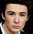 Pixelated close-up of a person's face with neutral expression, light skin tone, dark hair visible on left side, and white clothing collar.
