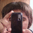 A person with brown hair holds a black smartphone in front of their face, obscuring the lower half of their face.