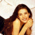 A pixelated image of a person with long dark hair wearing a light-colored sleeveless top, holding hands near the face in a static pose against a light background.