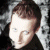 A pixelated image of a person with short brown hair wearing dark clothing, featuring partial text overlay on the face.