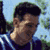 Pixelated static image of a person with dark hair wearing a blue jacket against an outdoor background with greenery and sky.