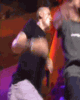 Animated sequence of two figures on a stage with red lighting, one in dark clothing holding a microphone and the other in gray shirt and light pants.