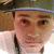 A pixelated close-up of a person wearing a dark cap with a light band, light-colored shirt, and dark tie, against a blurred background.