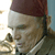Pixelated close-up of a person wearing a dark red fez-style hat and light-colored headband, with visible facial features and partial clothing.
