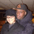 Two people in dark hooded coats and caps pose together under a ceiling, with one person's face partially obscured by light glare.