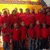 Group of people in red shirts standing in front of a yellow and brown building with a crowd behind them.
