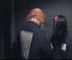 Two figures in dark clothing with light patches on their backs face away from the viewer, then turn to reveal masked faces and red hair.