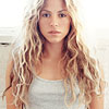 A person with long, wavy blonde hair wearing a light gray tank top against a bright, overexposed background.