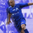 Pixelated static image of a person in blue athletic wear performing a soccer kick against a light purple background with white highlights.