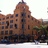 Pixelated image of a multi-story brick building with a rounded tower and dark windows, set against a light sky background.