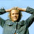 Pixelated static image of a person with light hair wearing a dark jacket, arms raised behind head against blue sky background.