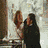 Two figures in a pixelated style, one with light hair and the other with dark hair, standing close together against a blurred background.