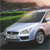 A silver sedan with a dark roof is positioned at a slight angle on a road, viewed from a low perspective with greenery and blurred background elements.