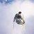 Pixelated static image of a skier in mid-air against a light blue sky background with faint cloud patterns.