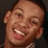 Pixelated close-up of a smiling person with short dark hair, visible ear, and light skin tone against a dark background.