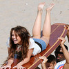 A woman with long brown hair lies on a striped surfboard, legs raised, wearing a white and blue top, held by multiple hands on a sandy beach.