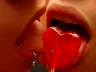 Close-up of a person's mouth holding a red heart-shaped object with visible teeth and lips, warm-toned skin tones, static image.