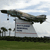 A pixelated image shows a large aircraft flying over a sign with horizontal stripes, set against a sky with clouds and palm trees in the background.