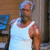 A man in a white tank top stands against a wooden background with blue fabric, his head slightly turned left.