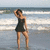 A woman in a black one-piece swimsuit stands on a beach with ocean waves behind her, her right hand raised to her head.