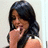 A woman with long dark hair in a sleeveless top, hand near mouth, smiling against light background.