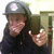 A person wearing a dark cap with a white circular emblem, dark clothing, and holding a dark rectangular object, with visible hand and background elements.