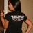 Pixelated image of a person in profile wearing black hooded clothing with white text on chest, holding an object near their face against a brown background.