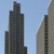 Pixelated image of two tall buildings against a light blue sky, with the left building taller and darker than the right one.